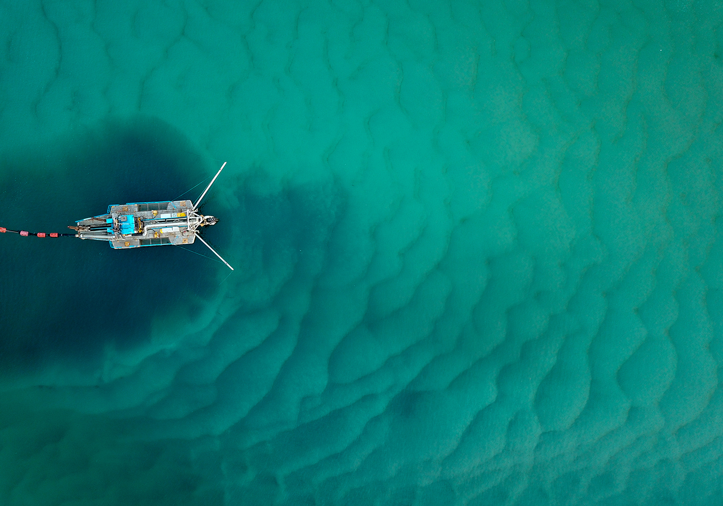 A view from above of a bottom trawler in the ocean. The dark water under the boat shows the destruction of the seafloor.