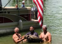 Three men holding a large catfish wast-deep in green water with a boat behind them flying the American flag.