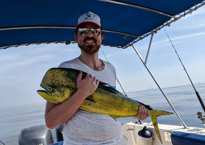 A man in a cap and sunglasses holding a Mahi Mahi fish on a boat