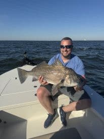 An angler proudly displaying a large Black Drum while seated on a boat, enjoying a sunny day on the water.
