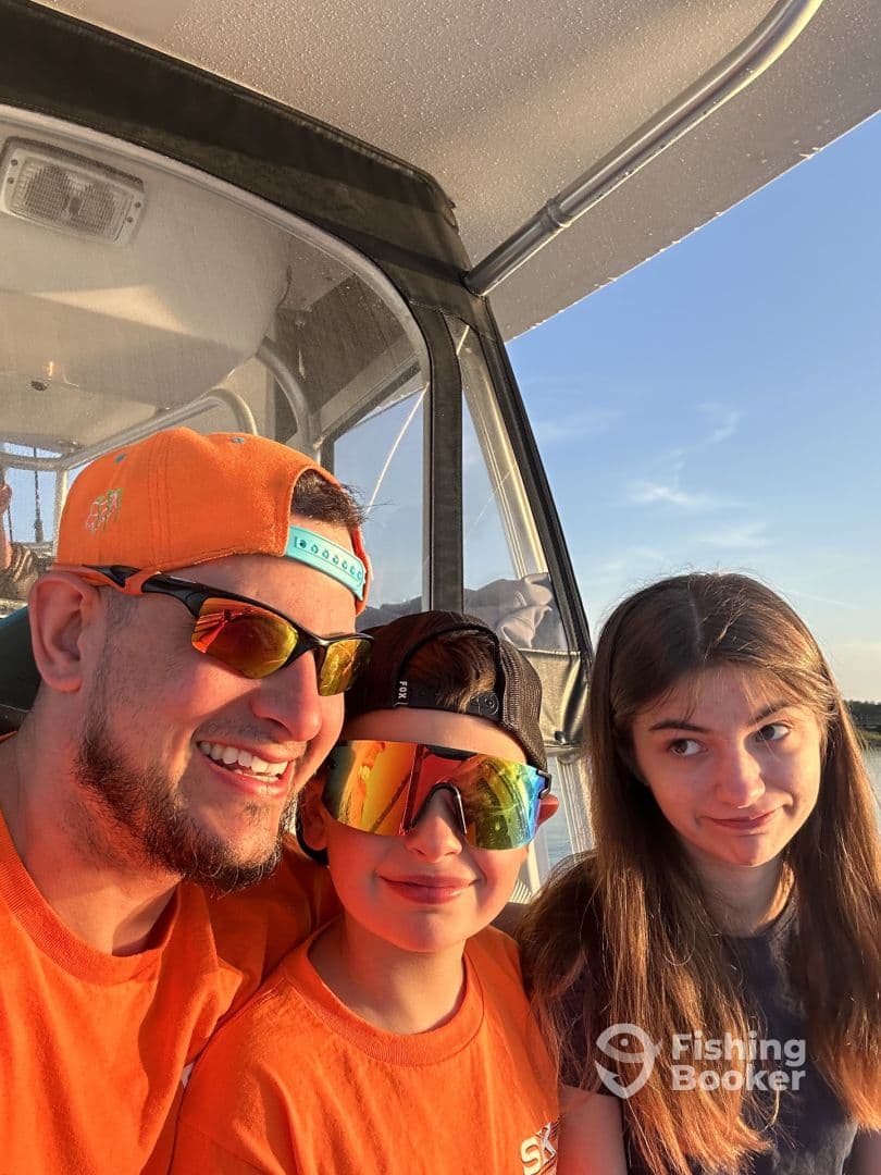 A family enjoying a sunset on a fishing boat, with two kids and an adult wearing matching orange shirts and sunglasses.