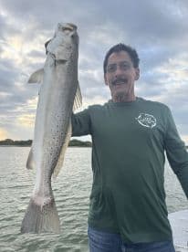 An angler proudly displaying a large Speckled Trout during a fishing trip, with a scenic water backdrop.