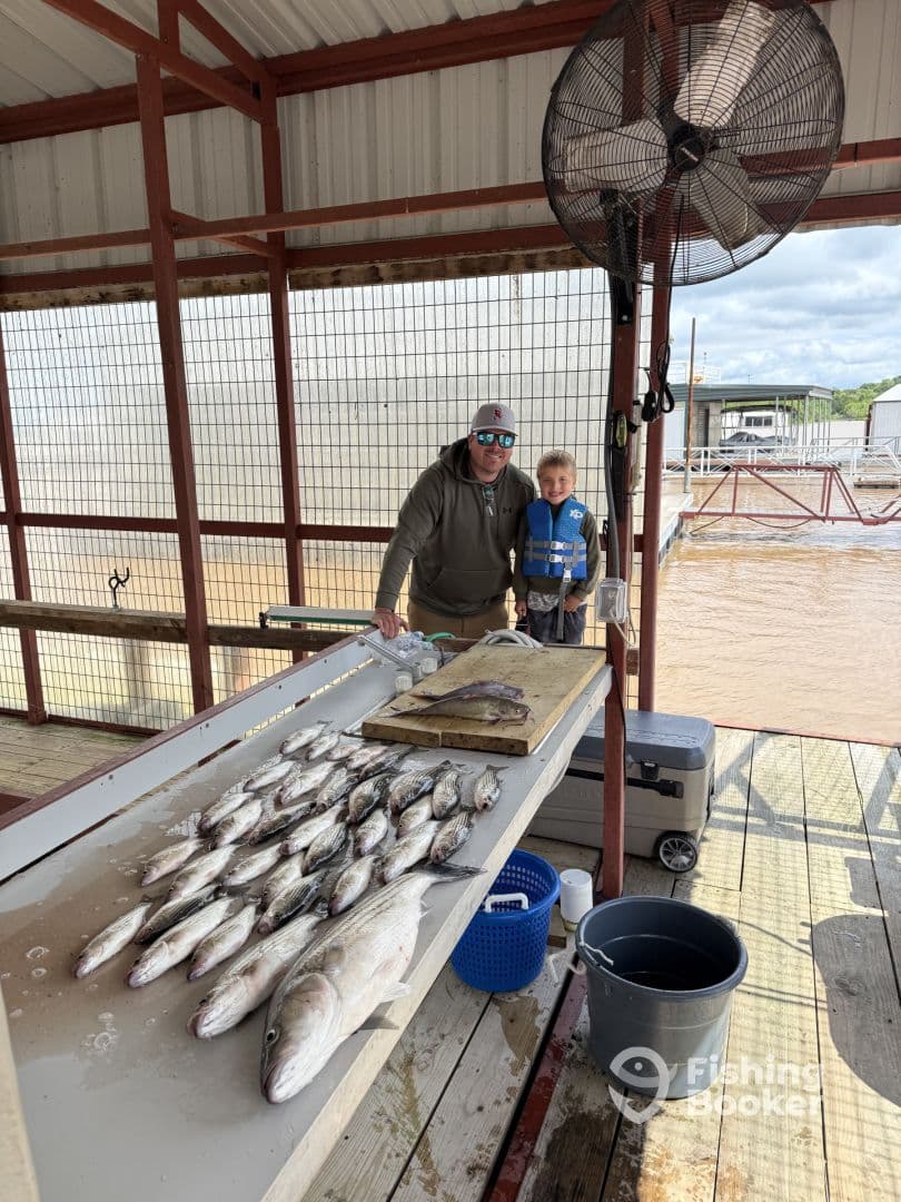 A father and son stand proudly beside a display of freshly caught fish on a cleaning table at a fishing dock, showcasing a successful day of fishing.
