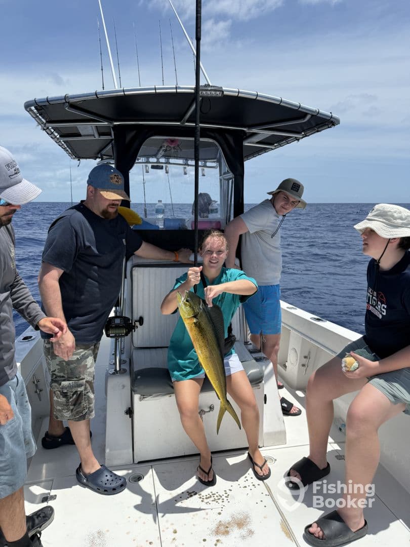 A young angler proudly displaying a Mahi Mahi aboard a fishing boat, surrounded by family members during a sunny day at sea.