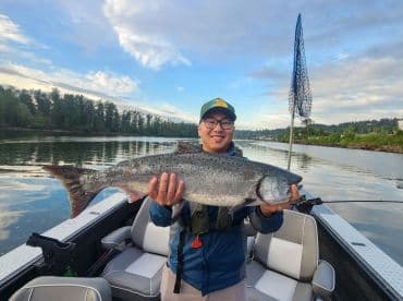 An angler proudly displaying a large Salmon while fishing from a boat on a calm river, showcasing a successful catch in a scenic environment.