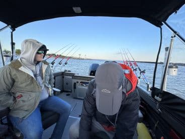 Two anglers seated inside a fishing boat, preparing for a day on the water with multiple fishing rods visible in the background.