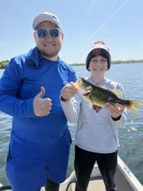 A young angler proudly displaying a large Largemouth Bass while fishing on a sunny day, accompanied by an adult on a boat.