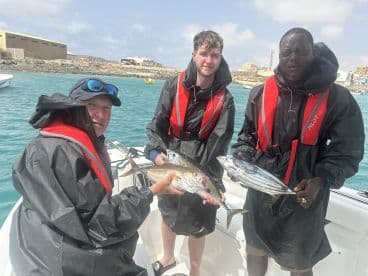 Three anglers proudly displaying their catch, including a large Yellowtail and a Bonito, while enjoying a fishing trip in a coastal setting.