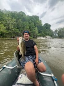 A young angler proudly displaying a large Largemouth Bass while fishing from a small boat in a scenic river setting.