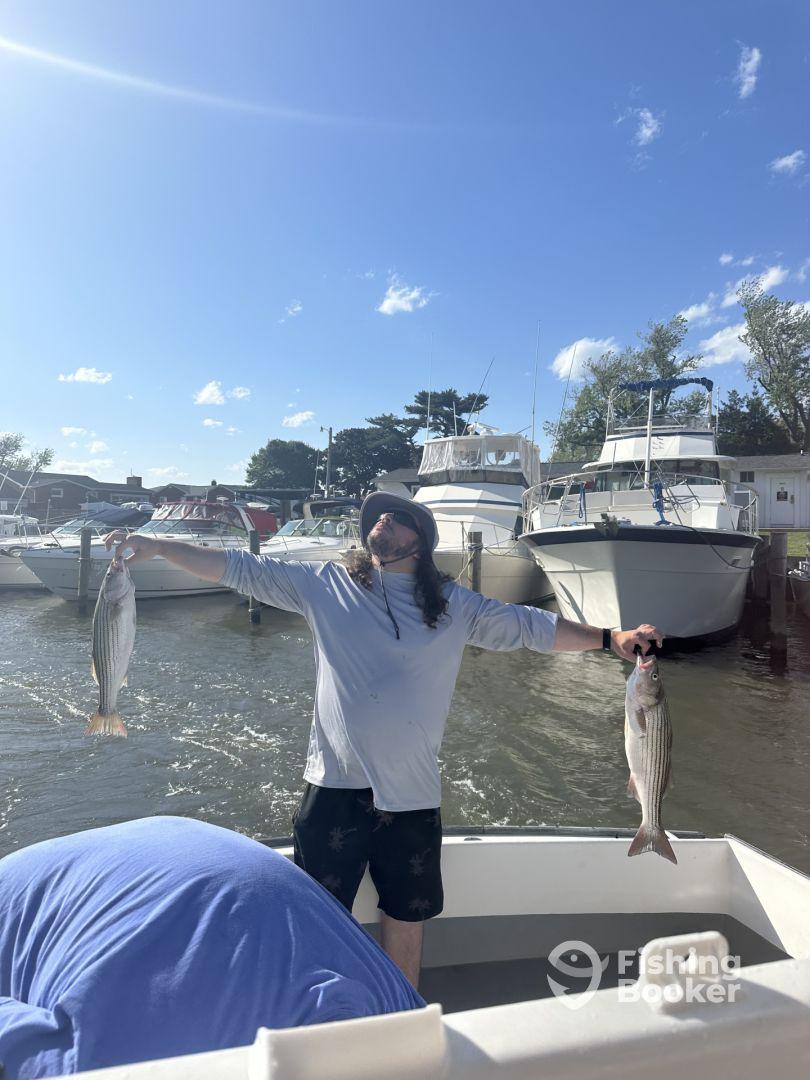 Angler proudly displaying two striped bass while on a fishing charter, with boats visible in the background.