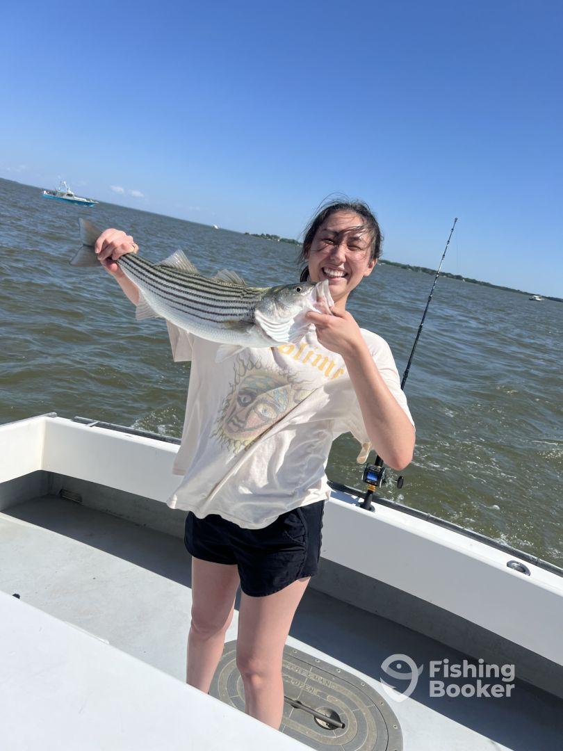 A young angler proudly displaying a striped bass while fishing on a boat in a sunny coastal environment.