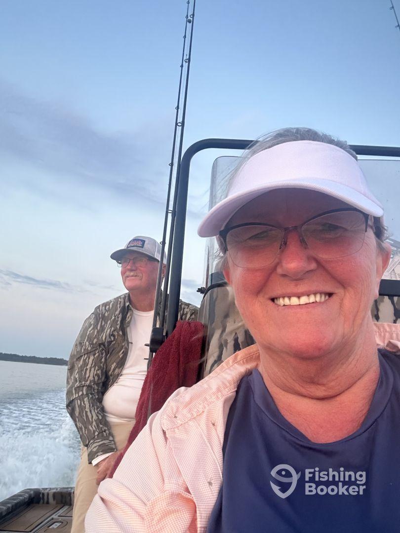 Two anglers enjoying a boat ride, with one taking a selfie while the other relaxes in the background. The setting suggests a peaceful fishing trip on calm waters.