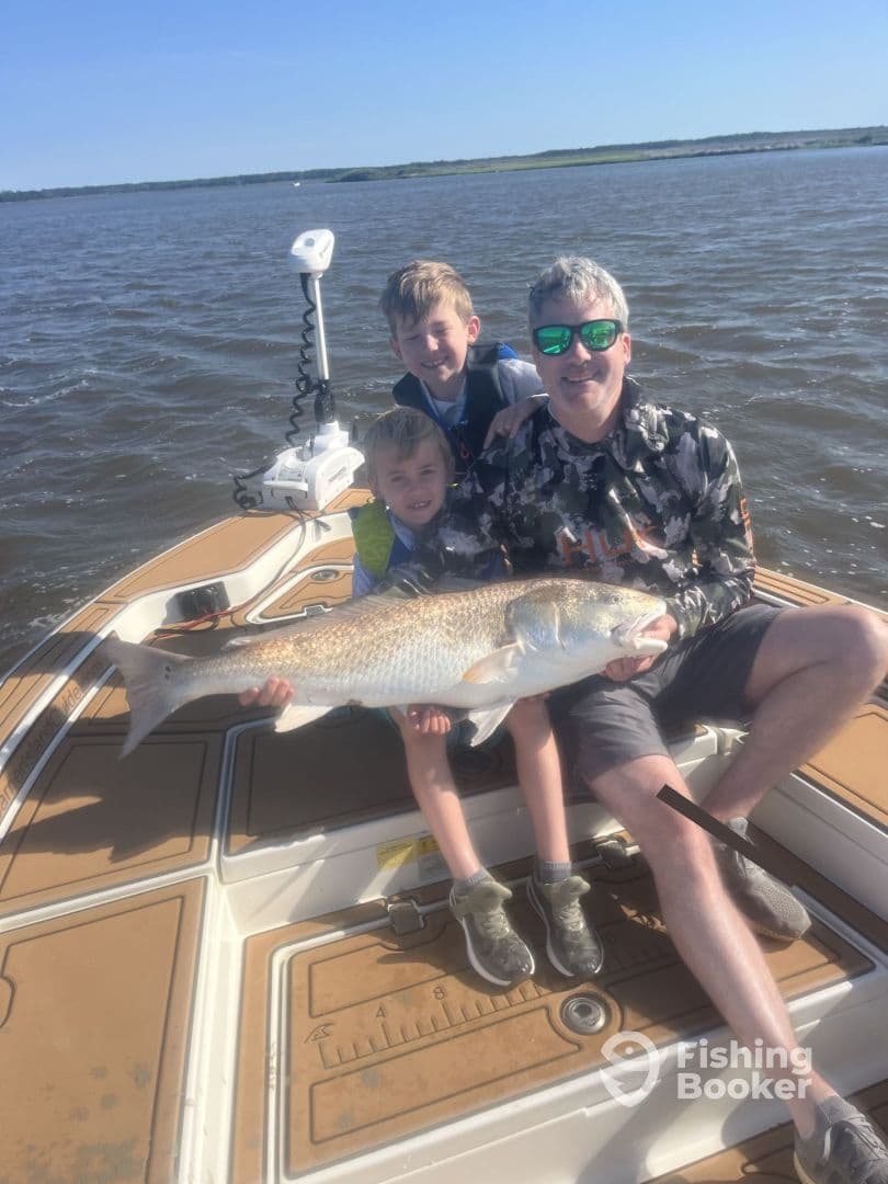 A family enjoying a successful fishing trip, proudly displaying a large Redfish on a boat in a scenic waterway.