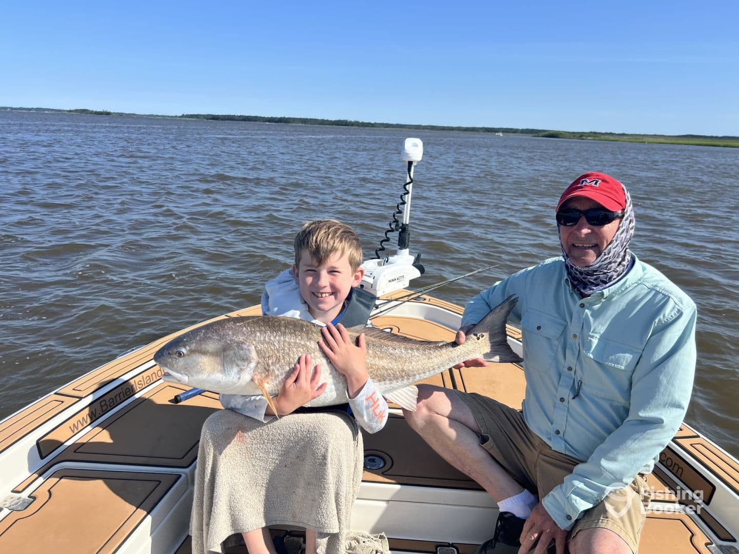 A young angler and an adult proudly displaying a large Redfish on a boat during a fishing trip.