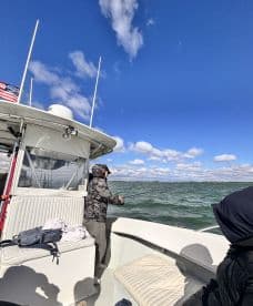 An angler casting a line from the deck of a fishing boat on a sunny day, with a clear blue sky and gentle waves in the background.