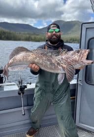 Angler proudly displaying a large Lingcod aboard a fishing boat in a scenic coastal environment.