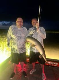 Two anglers proudly displaying a large fish, likely a Northern Pike, while fishing at night, showcasing their successful catch on a charter boat.