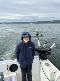 A young angler smiles while standing on a boat, with a fishing rod bent in action, indicating a catch in progress on a scenic waterway.