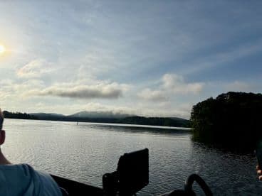 Scenic view from the side of a boat on a calm lake, surrounded by lush greenery and mountains in the background.