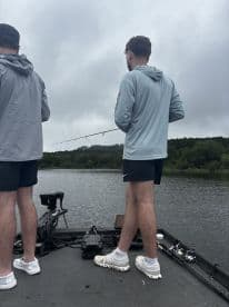 Two anglers casting lines from a bass boat on a cloudy day, surrounded by lush greenery.