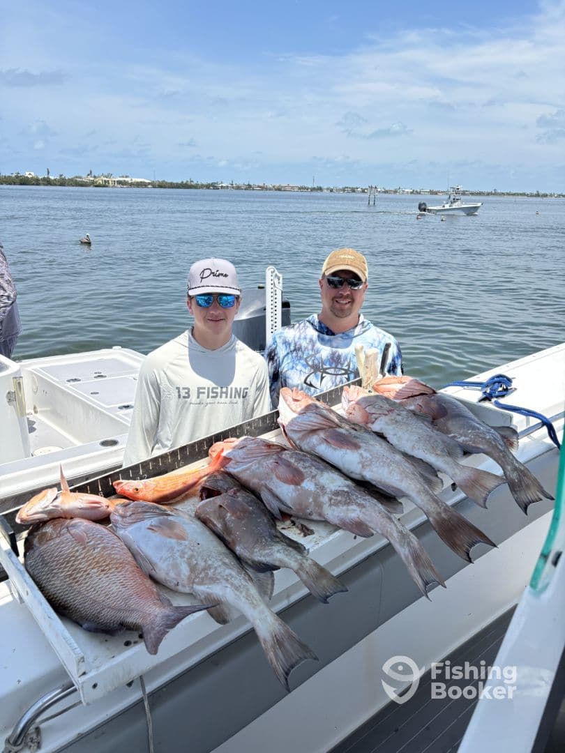 A young angler and an adult proudly pose with a variety of fish, including Snapper and Grouper, displayed on a catch board aboard a fishing boat.