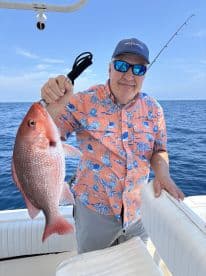 An angler proudly displaying a large Red Snapper while fishing offshore, showcasing a vibrant tropical shirt against a clear blue sky.