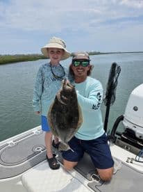 A young angler and an adult proudly display a large Flounder while fishing in a scenic coastal environment.