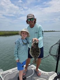 A young angler and an adult proudly displaying a large Flounder while fishing on a boat in a coastal waterway.