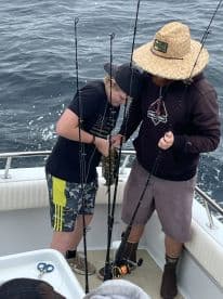 Two young anglers actively engaged in fishing on a boat, with one preparing to reel in a catch while the other assists.