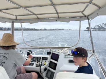 Two young anglers inside the helm of a boat, navigating through calm waters with a scenic coastal view.