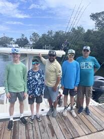 A group of four young anglers and an adult gather on the dock beside a fishing boat, showcasing their excitement for a day of fishing in a scenic coastal environment.