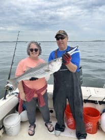 Two anglers proudly displaying a large Striped Bass while fishing from a boat, showcasing a successful day on the water.