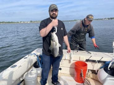 Angler proudly displaying a large Striped Bass on a fishing boat, with another crew member preparing gear in the background.