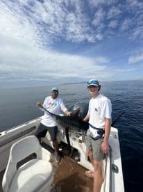 A young angler and a guide proudly displaying a large Marlin aboard a fishing boat in open waters.