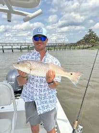 Angler proudly displaying a large Redfish while on a fishing charter, with a scenic backdrop of a pier and cloudy sky.