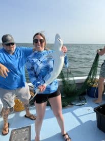 A proud angler displaying a large shark caught during a fishing trip, with fellow anglers on a boat in the background.