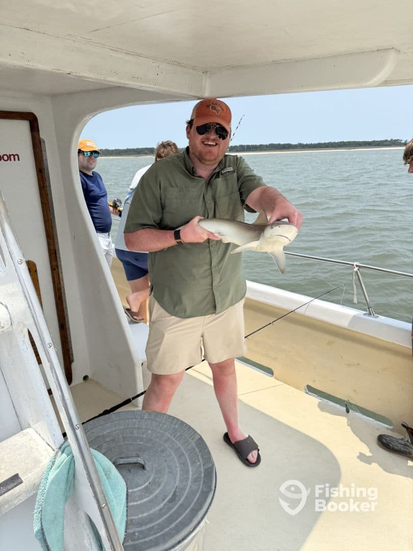 An angler proudly displaying a caught shark aboard a fishing charter, with a scenic water backdrop.