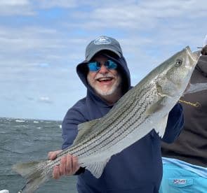An enthusiastic angler proudly displaying a large Striped Bass while fishing on a boat, showcasing a beautiful day on the water.