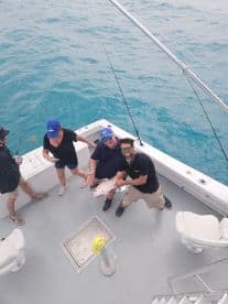 A group of anglers celebrating their catch on a charter boat, showcasing a large Snapper against the backdrop of clear blue waters.