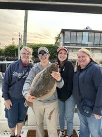 A group of four anglers proudly displaying a large Flounder on a fishing charter, showcasing a successful day on the water.