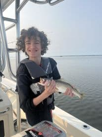 A young angler smiling while holding a Speckled Trout on a boat, showcasing a successful fishing trip.