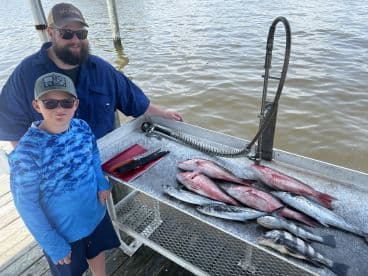 A father and son duo proudly display a variety of fish, including Redfish and Speckled Trout, on a cleaning table at the dock after a successful fishing trip.