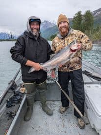 Two anglers proudly displaying a large Silver Salmon aboard a fishing boat in a scenic Alaskan backdrop.