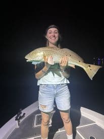 Angler proudly displaying a large Redfish while standing on the deck of a boat at night.