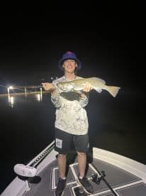A young angler proudly displaying a large Speckled Trout while fishing at night on a boat, illuminated by dock lights.