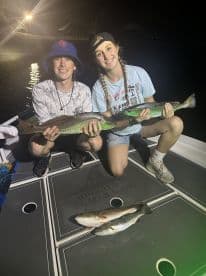 Two young anglers proudly displaying their catches, a pair of Speckled Trout, on a boat at night.