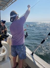 A young angler and an adult are actively fishing on a boat, with the adult reeling in a catch in sunny waters.