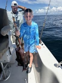 A young angler proudly displaying a large Grouper while fishing on a boat, showcasing a sunny day on the water.