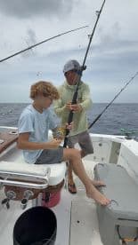 A young angler and an adult are engaged in fishing on a boat, with the young angler preparing to reel in a catch while the adult assists.