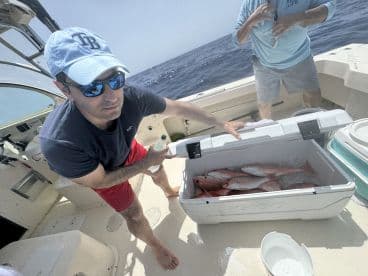 A fisherman on a boat is showing a cooler filled with freshly caught Red Snapper, highlighting a successful day of fishing in open waters.